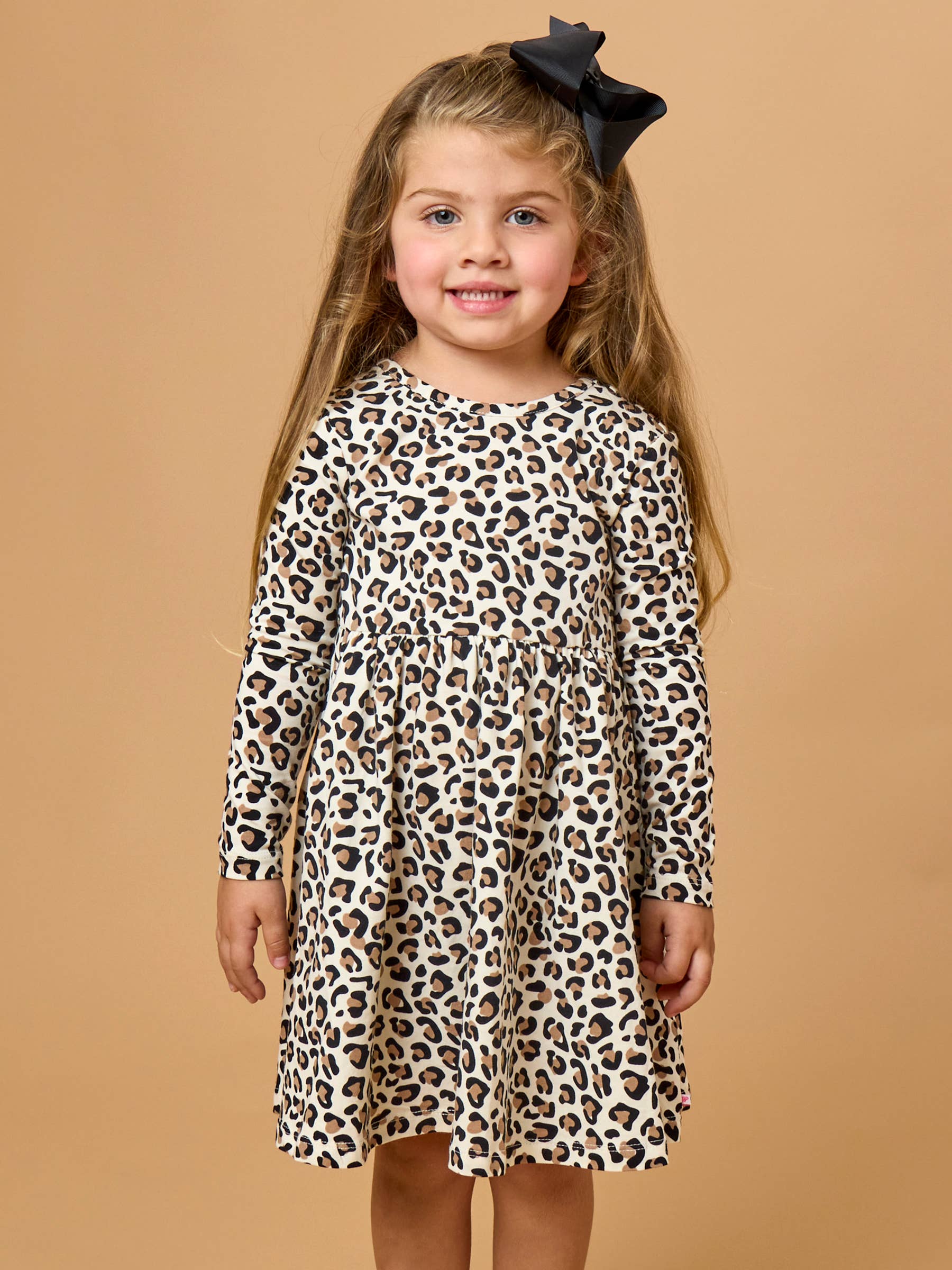 A young girl with long light brown hair wearing a long-sleeve leopard print dress and a black bow headband, standing against a solid tan studio background.