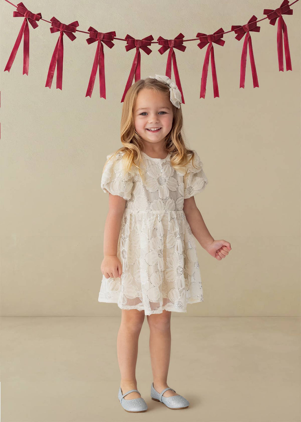Young girl in a beige lace dress standing in front of a beige wall with red ribbon decorations.