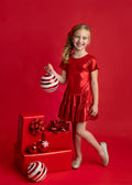 A young girl with blonde hair wearing a shiny red dress and holding a red and white striped ornament, standing next to a red gift box with bows and ornaments against a solid red background.