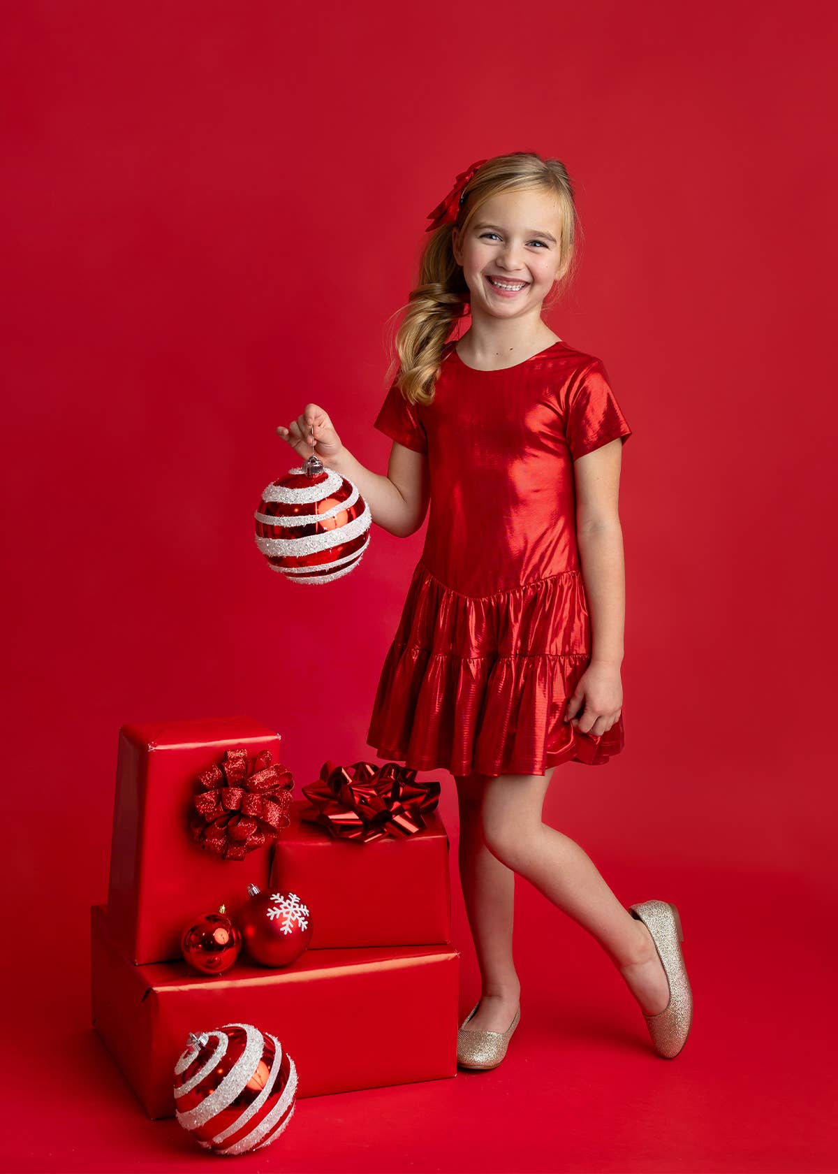 A young girl with blonde hair wearing a shiny red dress and holding a red and white striped ornament, standing next to a red gift box with bows and ornaments against a solid red background.