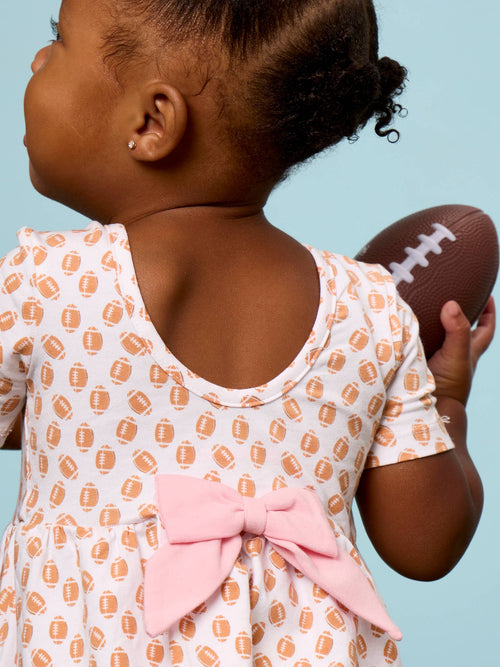 A toddler shown from the back wearing a white short-sleeve dress with an orange football print and a large pink bow at the back, holding a brown football against a light blue background.