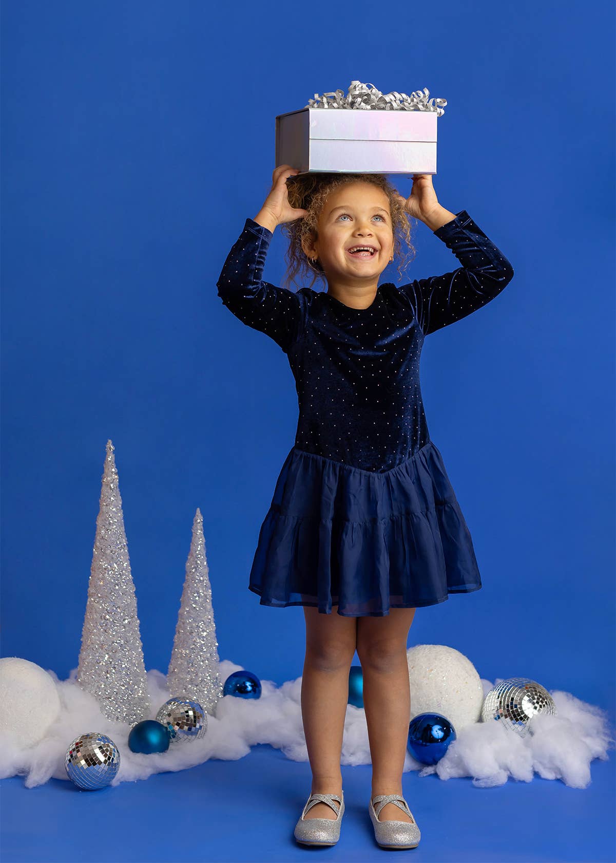 A young girl in a sparkling navy blue dress stands on a blue studio background, balancing a silver gift box on her head. Decorative white fluffy clouds, silver glitter trees, and blue and silver ornaments are scattered at her feet.