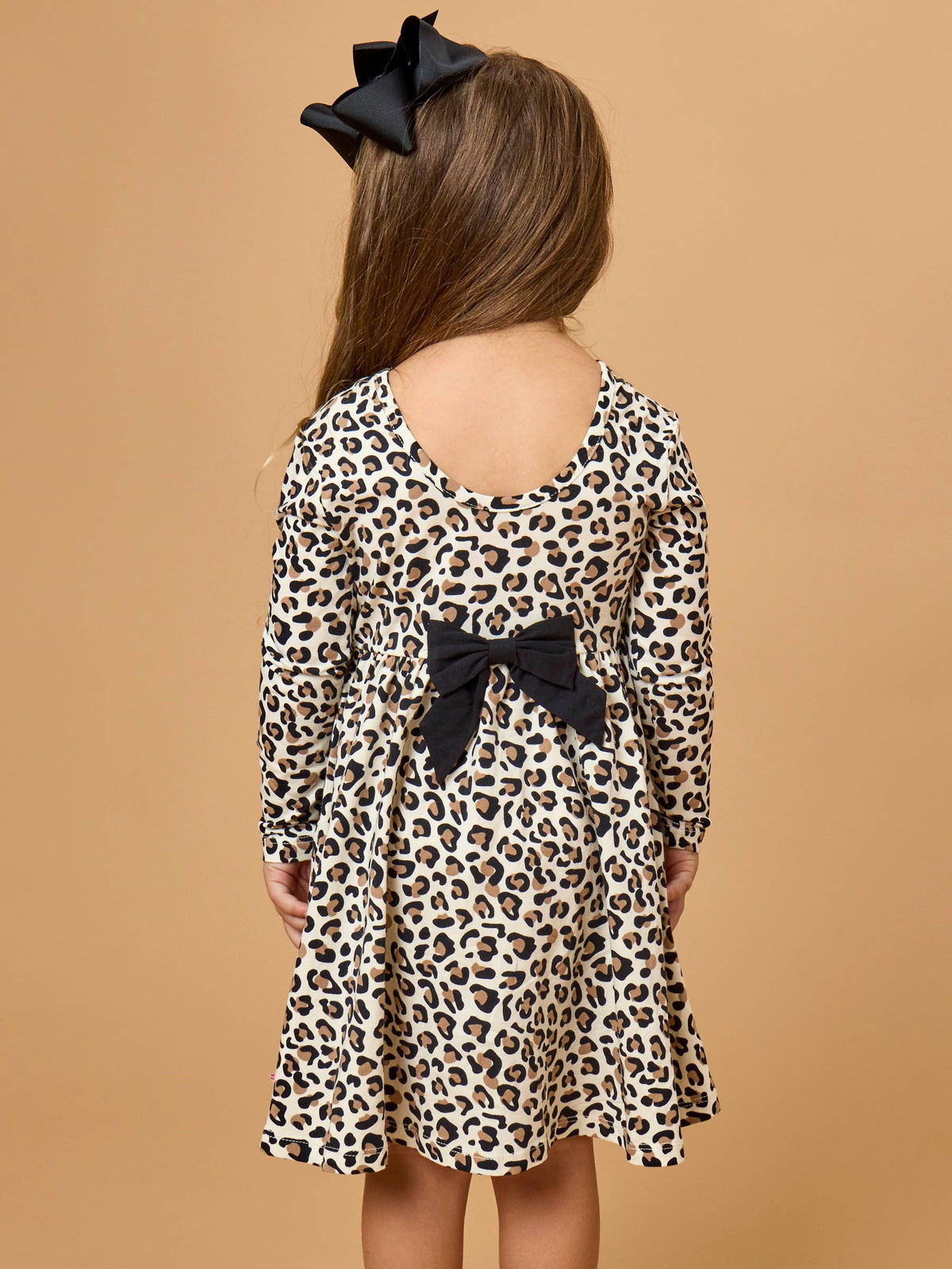 A young girl viewed from the back wearing a long-sleeve leopard-print dress with a black bow at the back waist. She has a large black hair bow in her hair and is standing against a solid tan background.