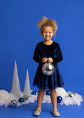 A young girl with curly blonde hair stands against a solid blue studio background, wearing a dark navy blue long-sleeve dress with a sparkly bodice and a tulle skirt. She is holding a silver disco ball ornament.