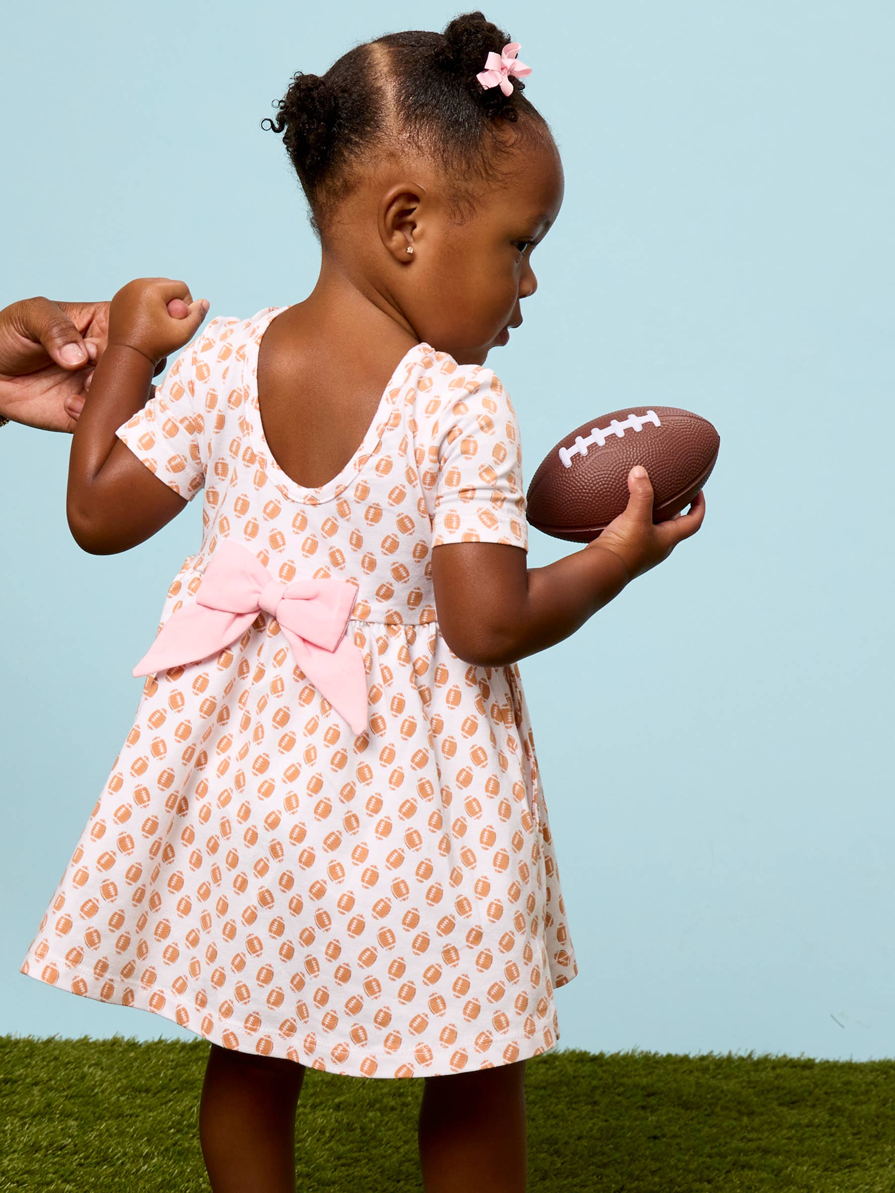 A toddler girl wearing a short-sleeve dress with a peach and white pattern, a large pink bow at the back, and holding a football, standing on artificial grass against a light blue background.