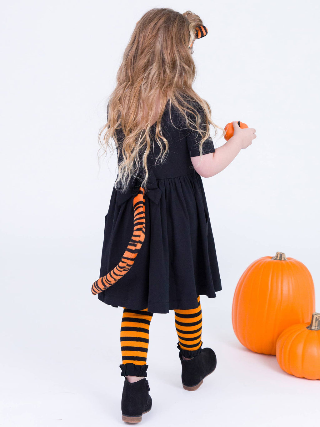 A young girl with long blonde hair is shown from behind wearing a black dress with an orange and black tiger-striped tail accessory, striped tights, and black boots. She is holding a small orange pumpkin next to two larger pumpkins on a white studio background.