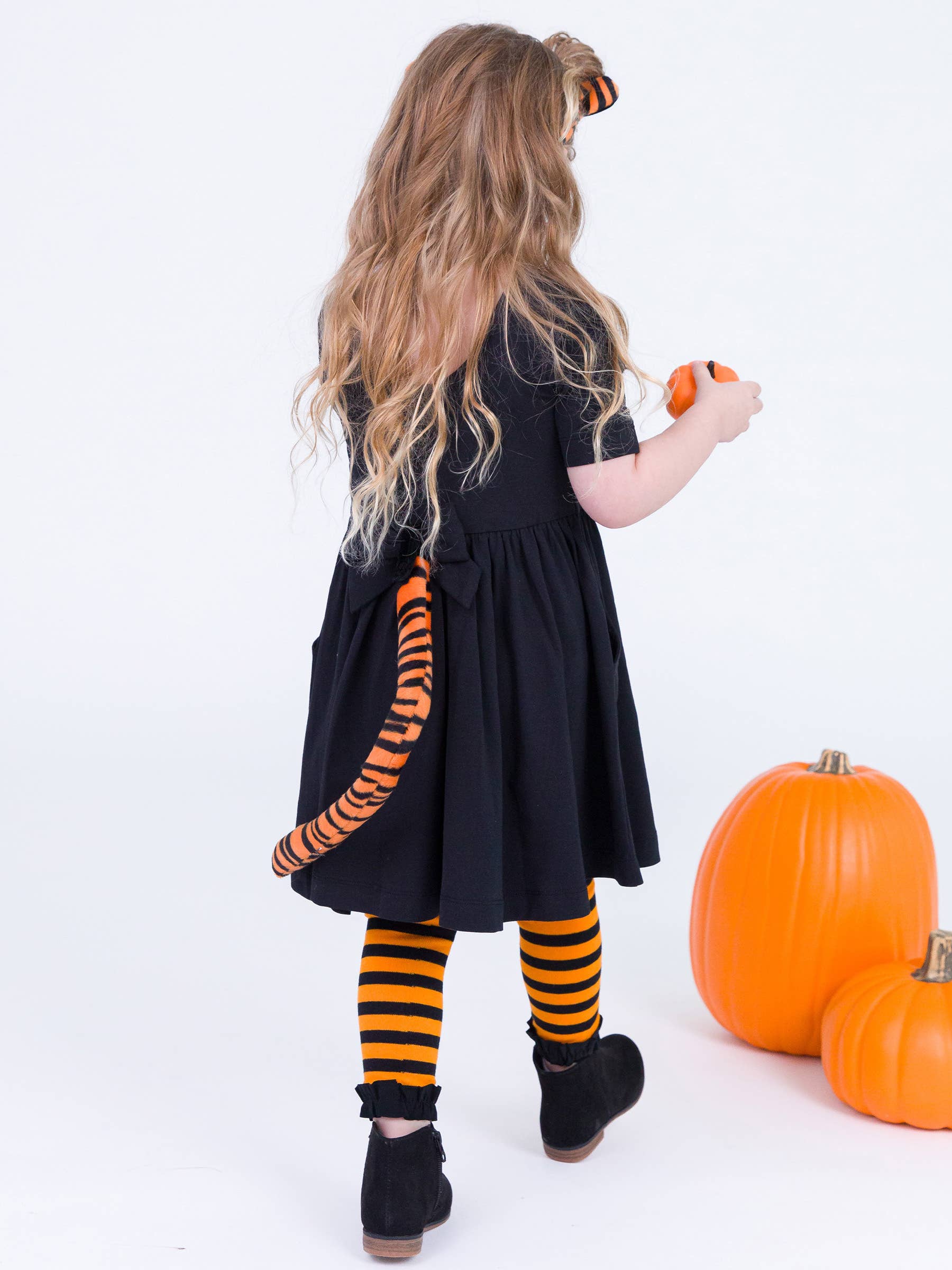 A young girl with long blonde hair is shown from behind wearing a black dress with an orange and black tiger-striped tail accessory, striped tights, and black boots. She is holding a small orange pumpkin next to two larger pumpkins on a white studio background.