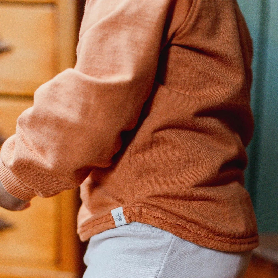 Close-up of a child wearing a rust-orange long-sleeve organic cotton sweatshirt and light gray pants, with a small white brand tag visible near the hem.