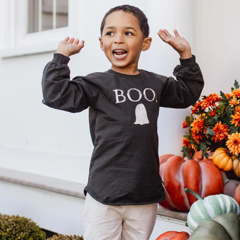 Child wearing a black 'BOO' organic cotton sweatshirt with a ghost graphic, standing in front of pumpkins and flowers.