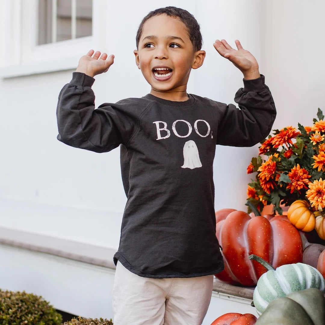 Child wearing a black 'BOO' organic cotton sweatshirt with a ghost graphic, standing in front of pumpkins and flowers.
