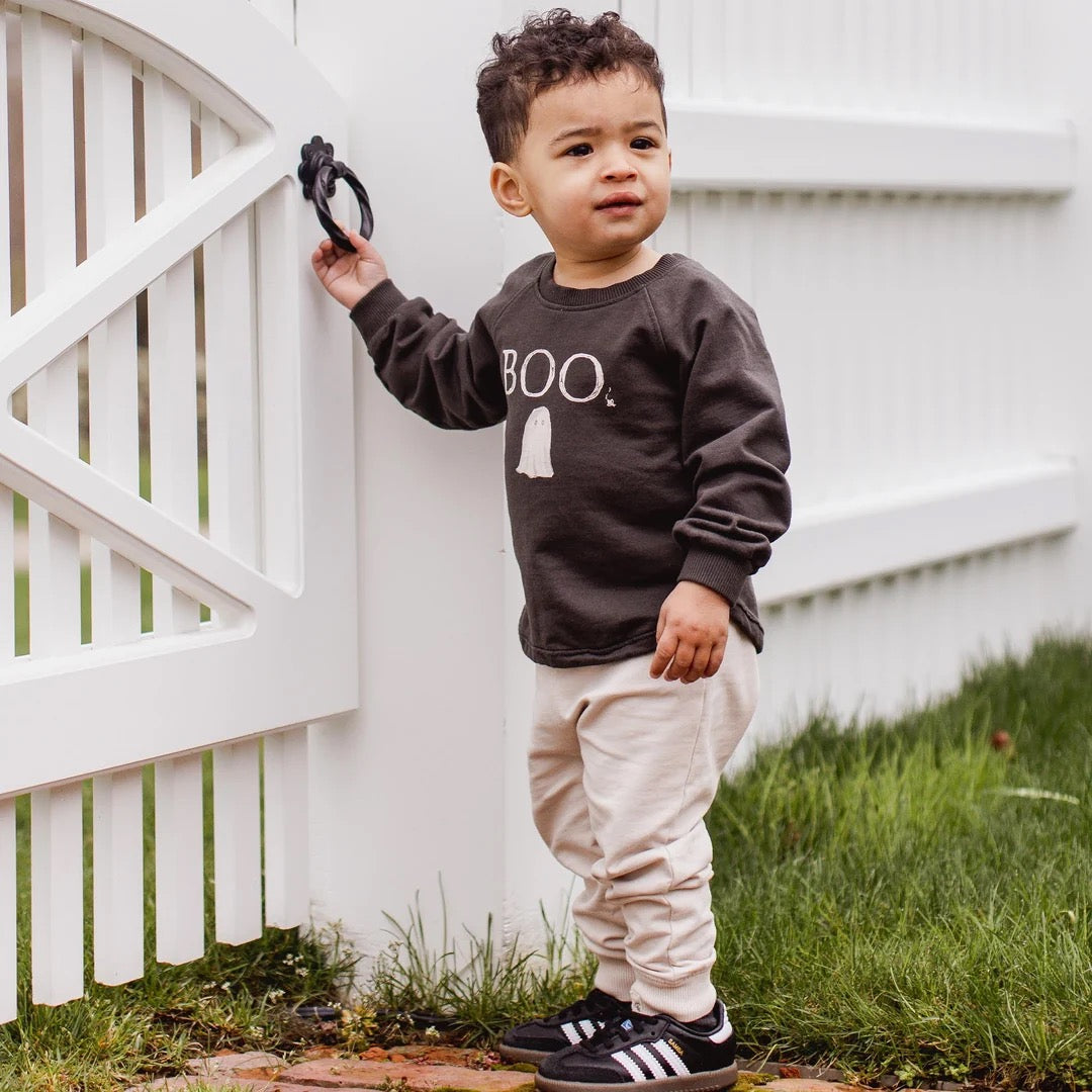 Child wearing a black organic cotton sweatshirt with 'BOO' printed on it, standing in front of a white picket fence.