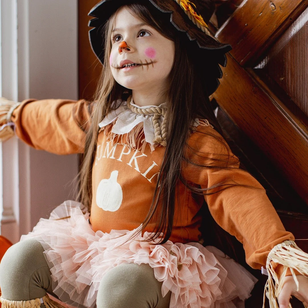 A young girl wearing a long sleeve organic cotton sweatshirt featuring an orange long-sleeve top with 'PUMPKIN' and a pumpkin graphic, a light pink ruffled tutu skirt, olive green leggings, and a black straw hat, sitting in front of a wooden door.