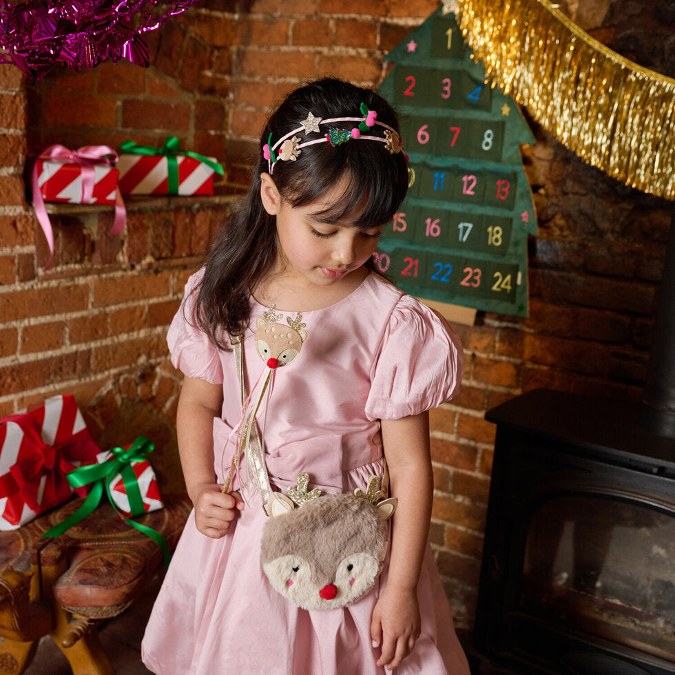 Young girl in a pink dress holding a reindeer bag in front of a Christmas tree and presents.