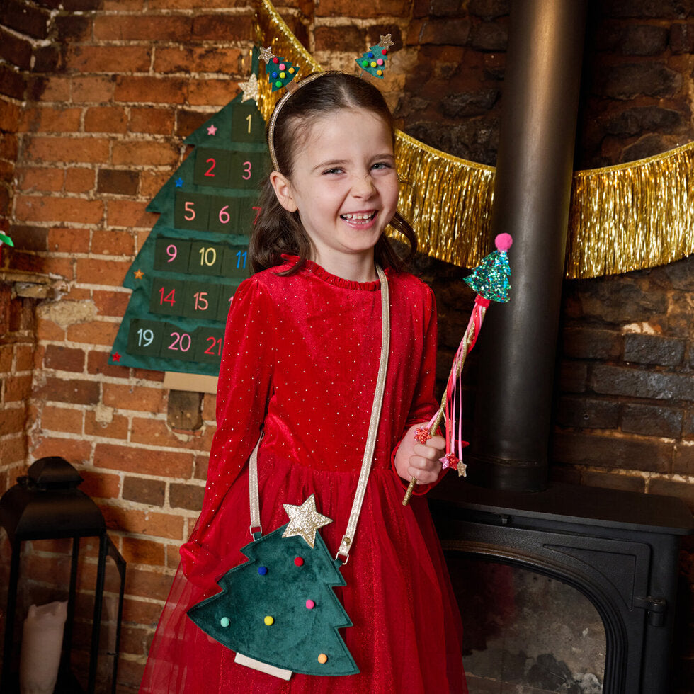 A young girl smiling in a red dress, wearing a green Christmas tree crossbody bag with a star and holding a wand in front of a brick fireplace with gold garland.
