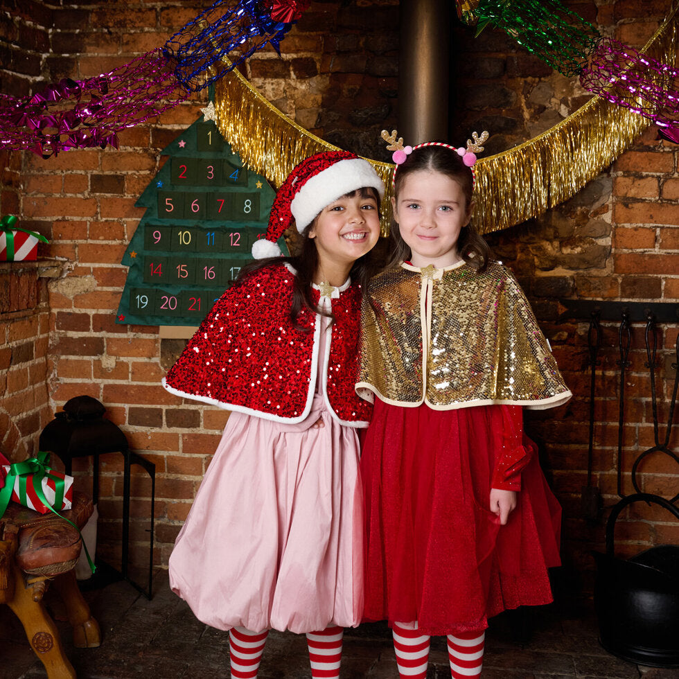 Two young girls wearing festive Christmas costumes posing in front of a rustic brick fireplace. One girl wears a Santa hat and a red sequined cape over a light pink dress, while the other wears a gold sequined cape over a red dress.