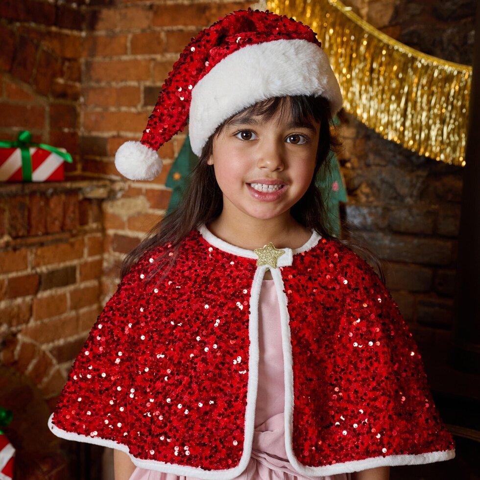 Young girl wearing a red sequin cape and Santa hat in front of a brick wall with Christmas decorations.