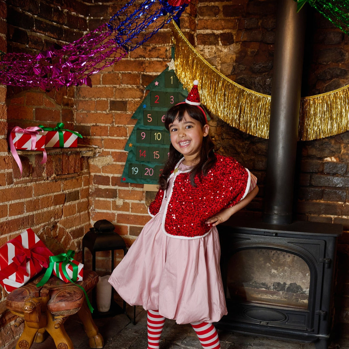 Young girl in festive outfit standing in front of a brick wall with Christmas decorations.