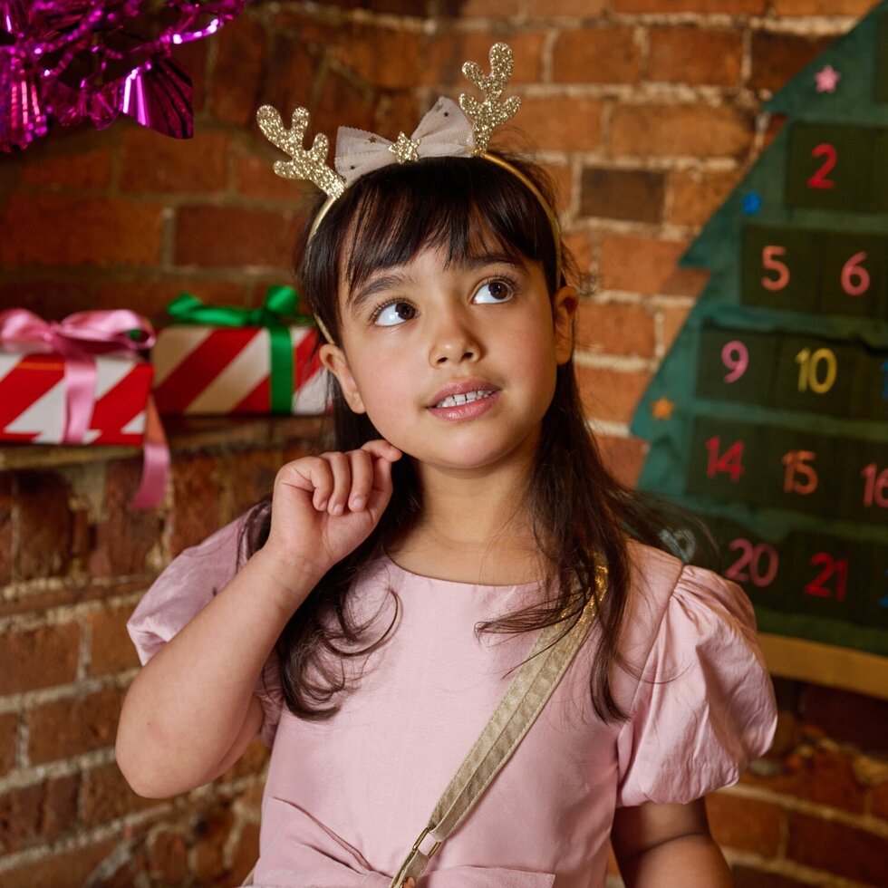 A young girl with dark hair wearing a pink dress and a gold glitter reindeer antler headband, looking up and to the side in a festive setting with a brick wall background.