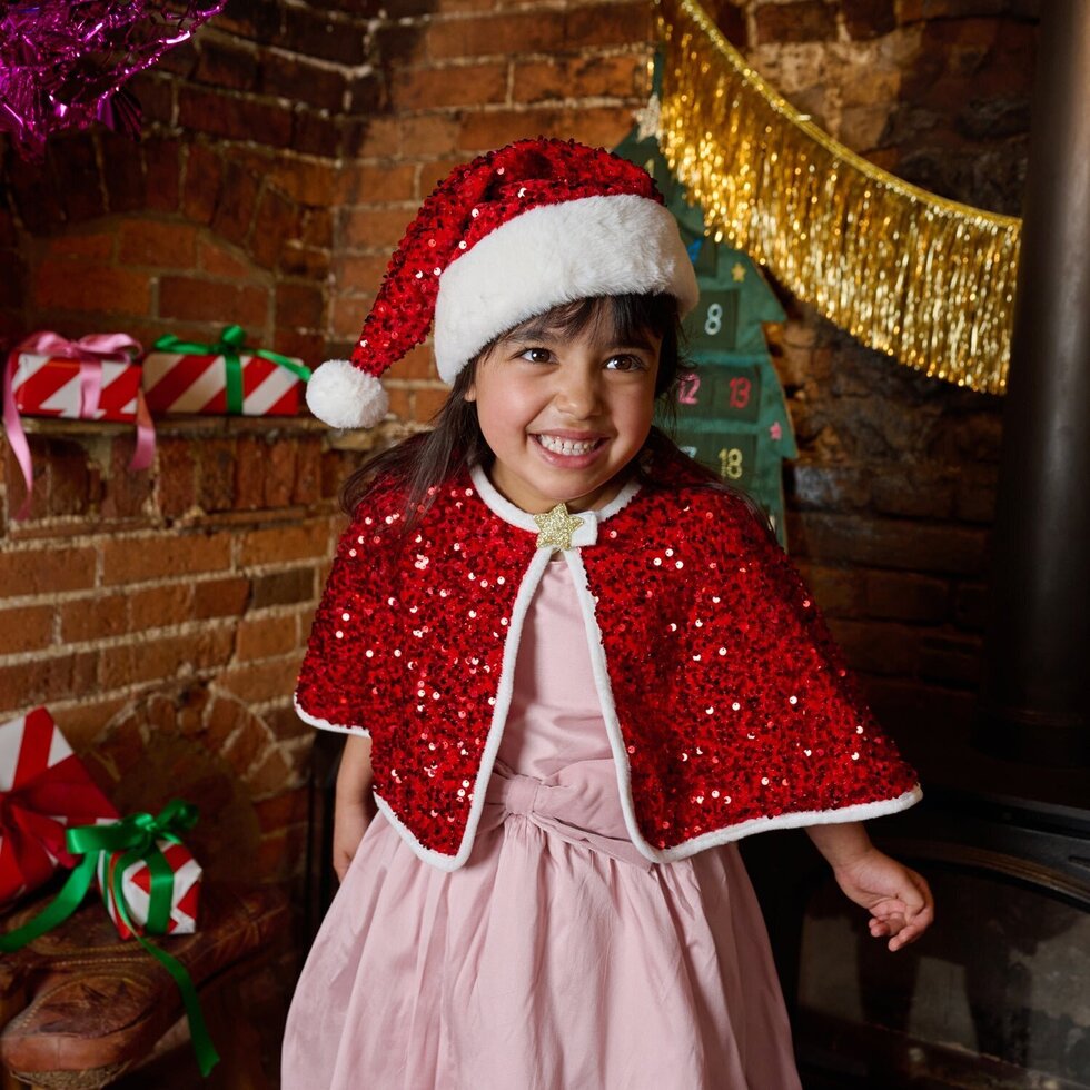 A young girl smiling in a festive holiday setting, wearing a red sequined Santa hat with white trim and a matching red sequined cape with white trim over a light pink dress. The background features a rustic brick fireplace with holiday decorations, including a gold tinsel garland and wrapped gifts.