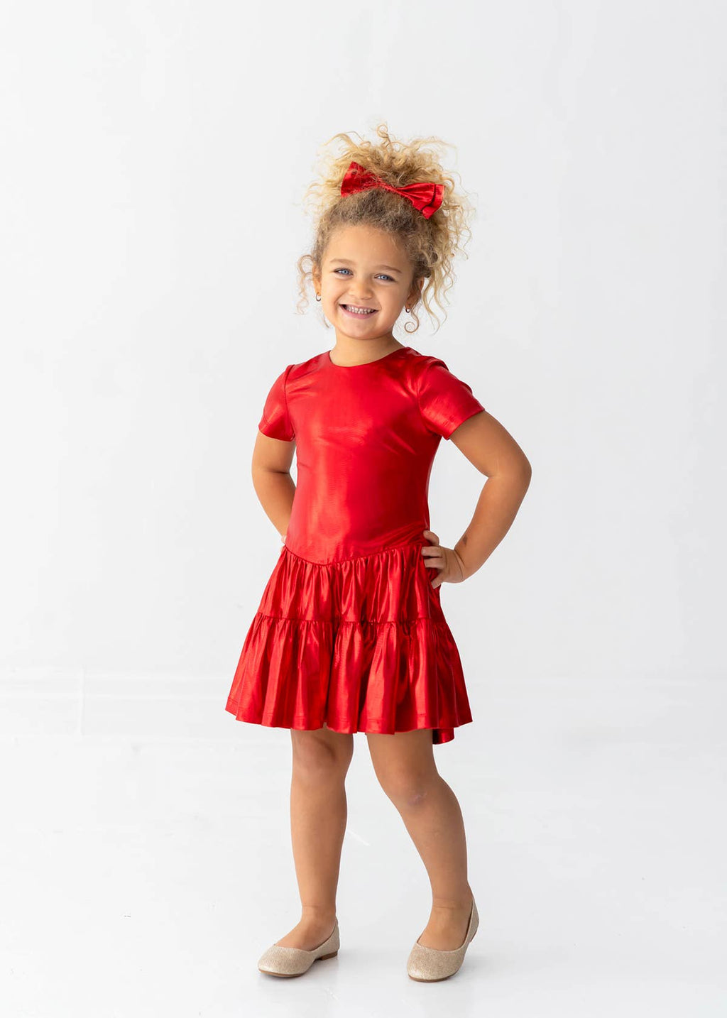 A young girl with curly hair wearing a shiny red short-sleeve dress and beige shoes, standing against a plain white studio background.