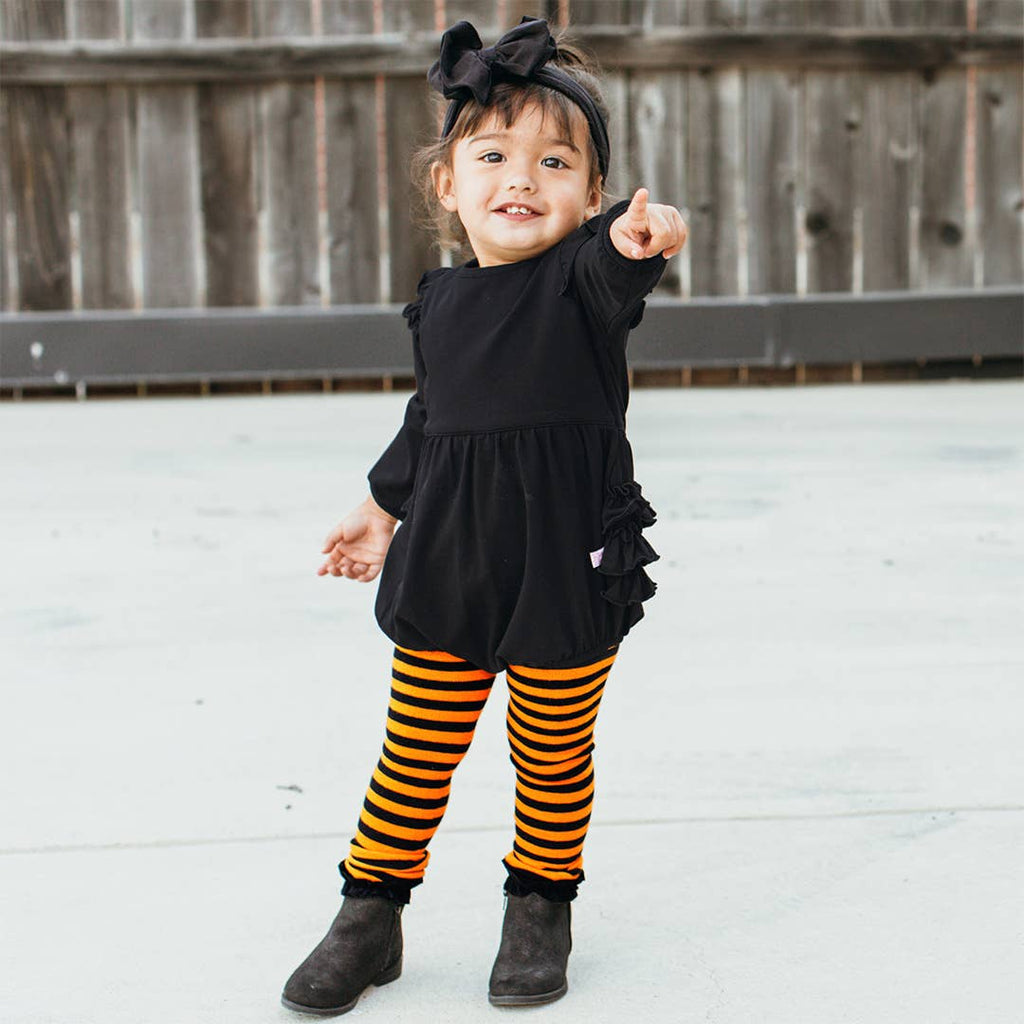 A toddler girl wearing a black long-sleeve dress with ruffled trim, black and orange striped tights, and black boots, standing outdoors in front of a wooden fence.