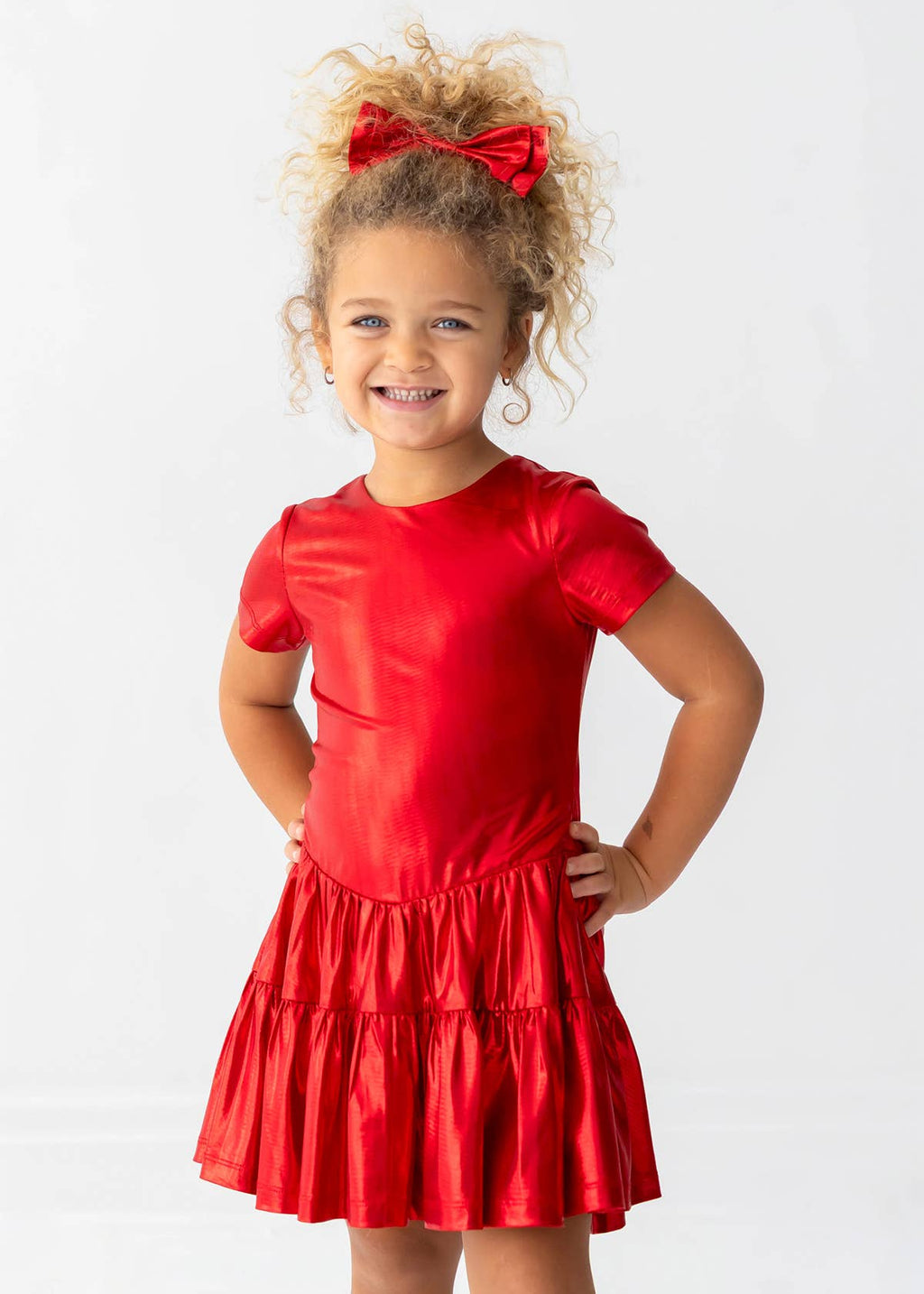 A young girl with curly blonde hair wearing a shiny red short-sleeve dress with a tiered skirt and a matching red bow in her hair, posing against a white studio background.