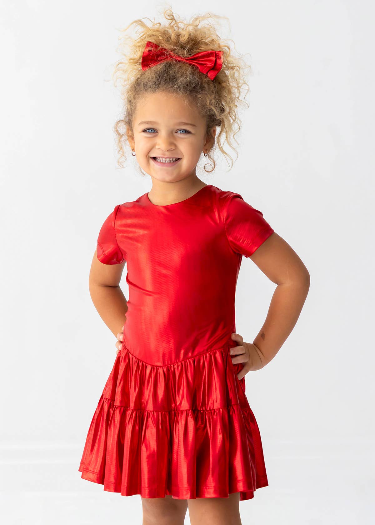 A young girl with curly blonde hair wearing a shiny red short-sleeve dress with a tiered skirt and a matching red bow in her hair, posing against a white studio background.
