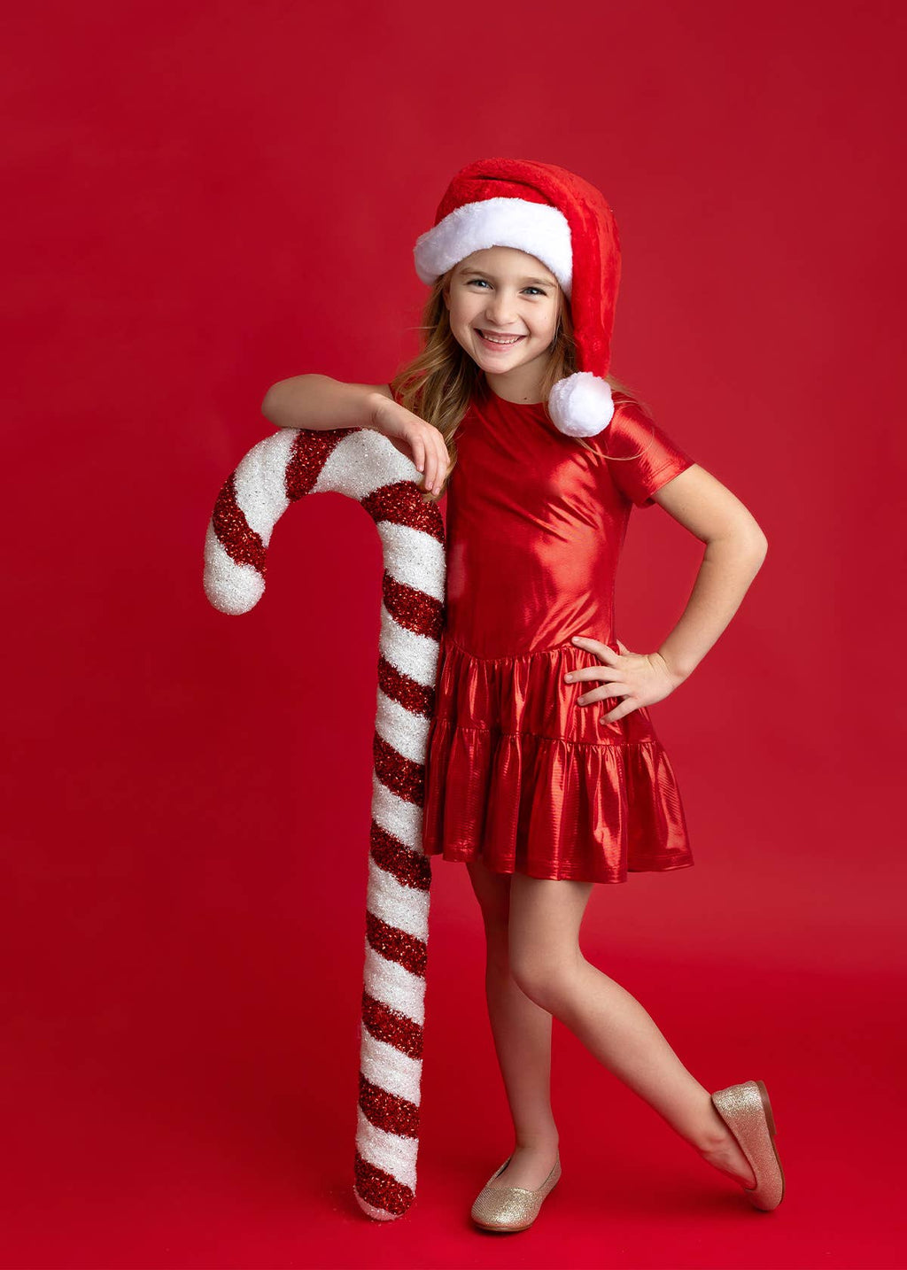 A young girl posing in a shiny red Christmas dress and Santa hat, holding a large red and white striped candy cane prop against a solid red studio background.