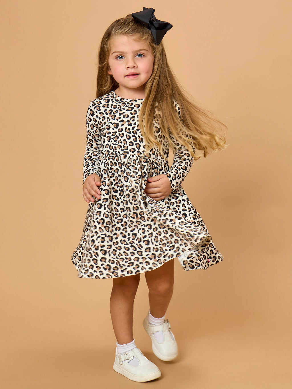 A young girl with long blonde hair wearing a long-sleeve leopard print dress, white sneakers, and a black bow headband, standing against a solid beige studio background.