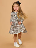 A young girl with long blonde hair wearing a long-sleeve leopard print dress, white sneakers, and a black bow headband, standing against a solid beige studio background.