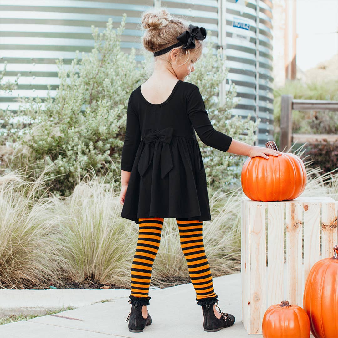 A young girl wearing a black long-sleeve dress with a bow at the back, black and orange striped tights, and black shoes, standing outdoors next to pumpkins.