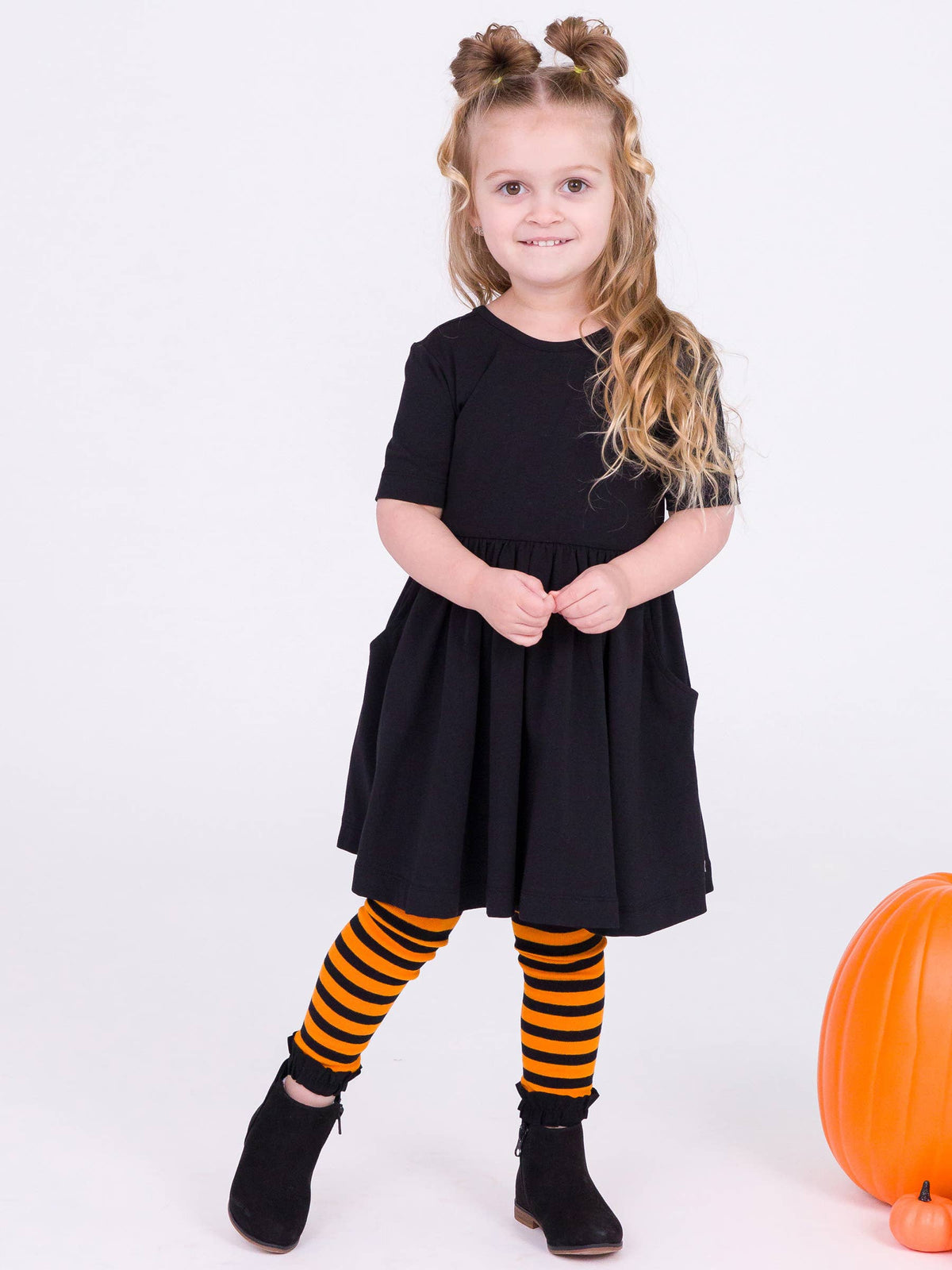 A young girl with blonde hair in two buns stands against a white studio background wearing a black short-sleeve dress, black and orange striped tights, and black ankle boots. An orange pumpkin is partially visible at the bottom right.